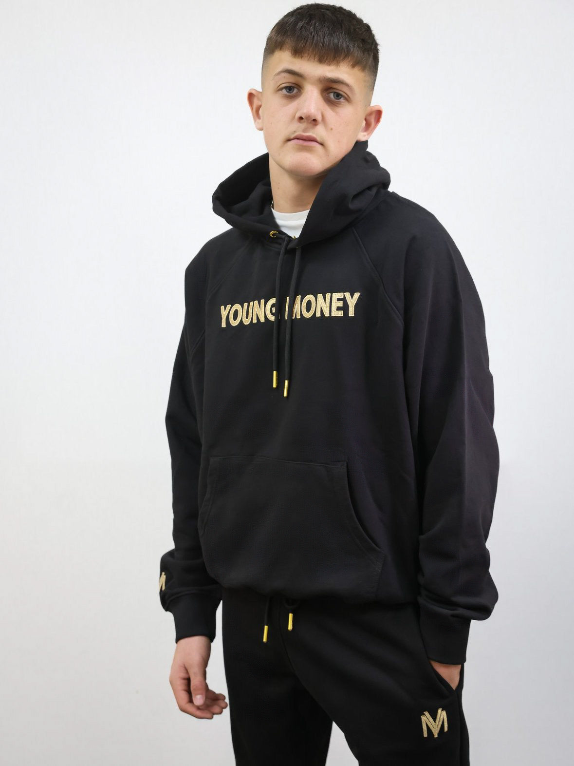 Black organic cotton oversized hoodie with gold embroidered young money lettering on the front and a gold embroidered YM logo on the sleeve on a male model size small featuring matching black and gold joggers